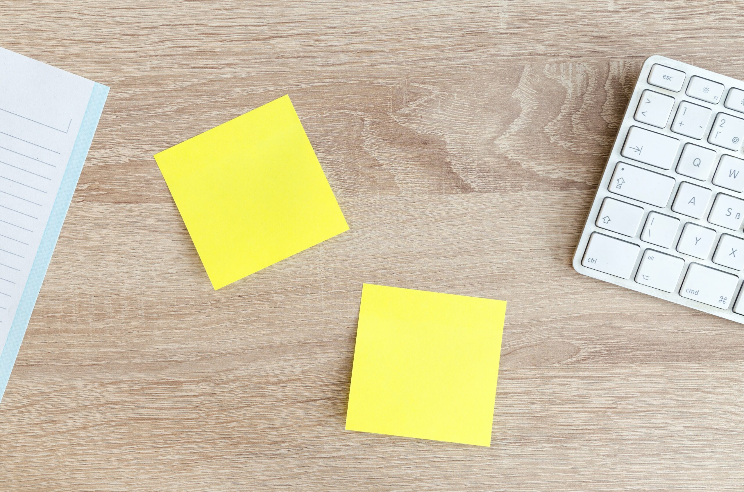 Two yellow sticky notes on a wooden table. There is a white keyboard that can be partially seen on the right side and a white notepad on the left side.