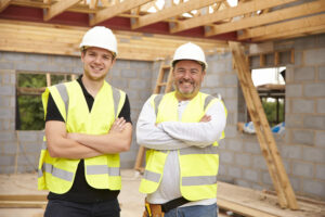 Younger male tradie standing at a construction site with an older male tradie. They are both wearing hard hats and a high vis vest with their arms crossed smiling.