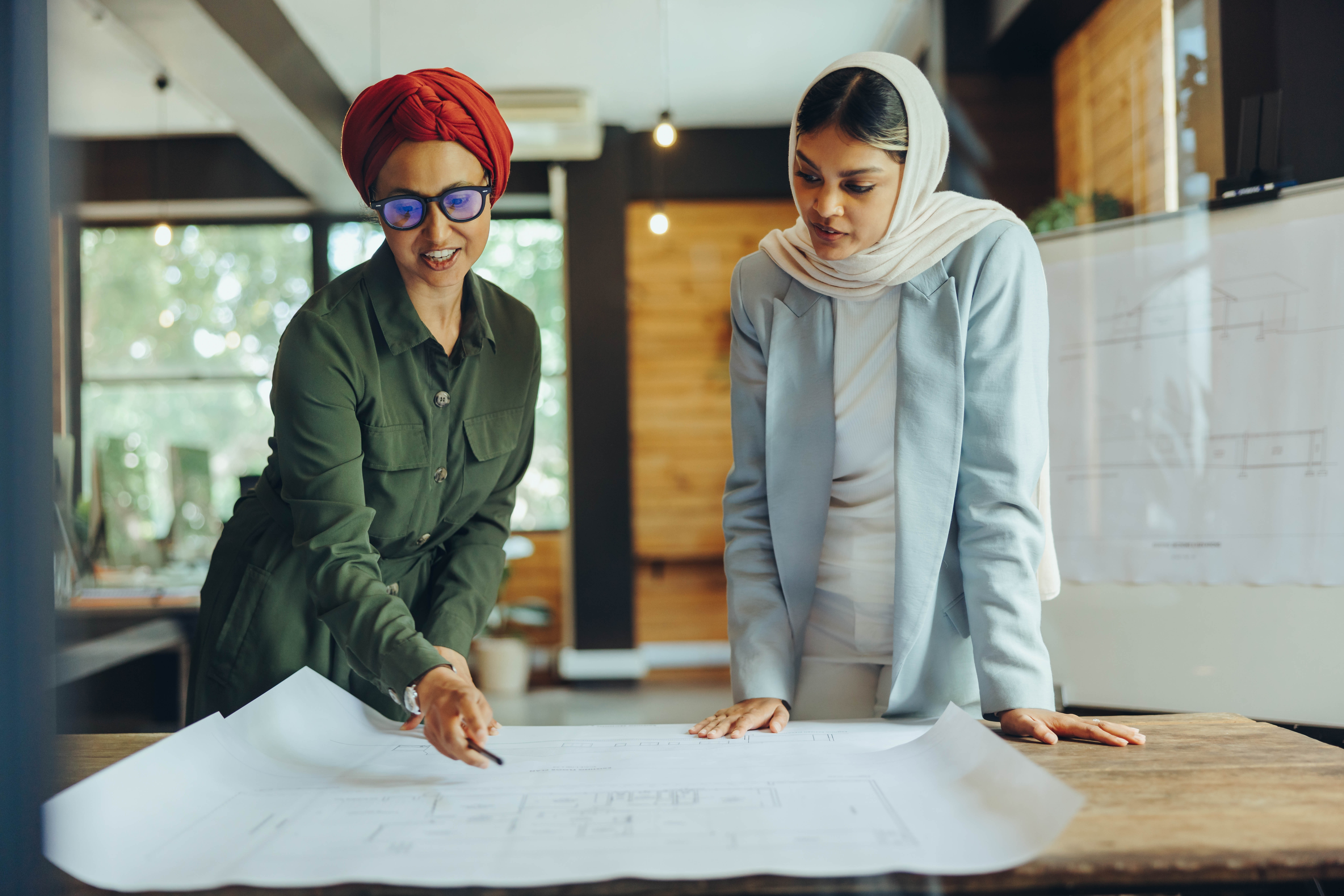 Two women wearing cultural head pieces standing over a desk. They have a large poster in front of them and are discussing the plans on the poster.