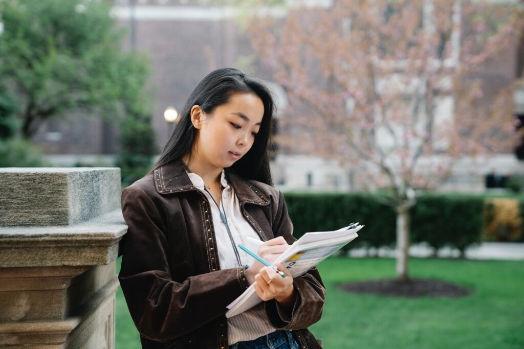 Young asian woman writing notes in her journal while leaning against a concrete pillar outdoors