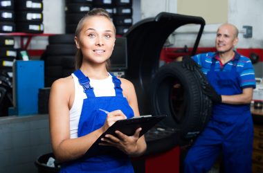 Young happy russian female technician in overalls holding clipboard in car workshop