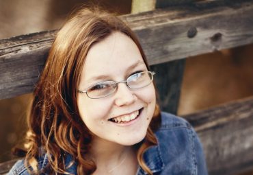 Young girl wearing glasses and a denim jacket is smiling looking at the camera. She has short golden brown hair.