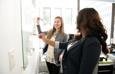Two female coworkers holding markers and writing on a whiteboard