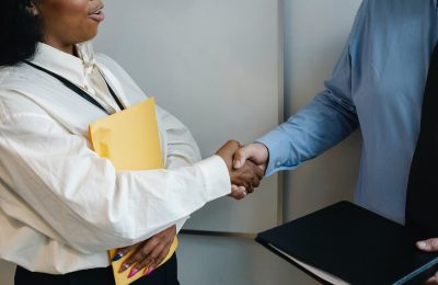 Female worker shaking the hand of her male coworker. They are both wearing dress shirts and have a folder in their free hand.
